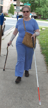 Photo shows the woman walking along a sidewalk, supporting herself with a support cane in her right hand, and reaching a long white cane ahead of her with her left hand (her right foot is forward, and the tip of the long cane is far in front of her left foot).